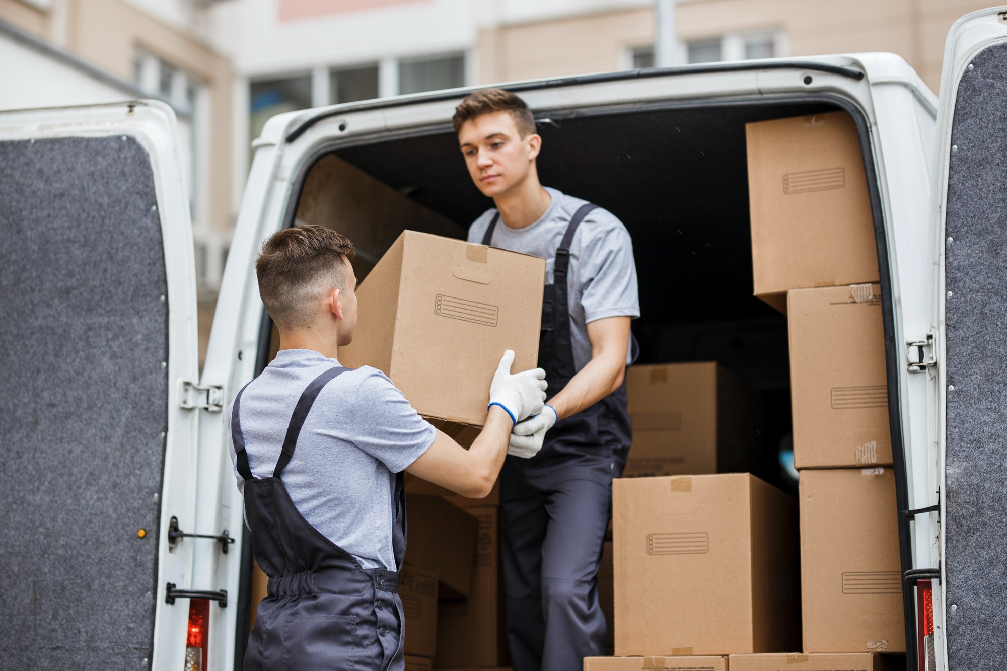 Two young handsome movers wearing uniforms are unloading the van full of boxes. House move, mover